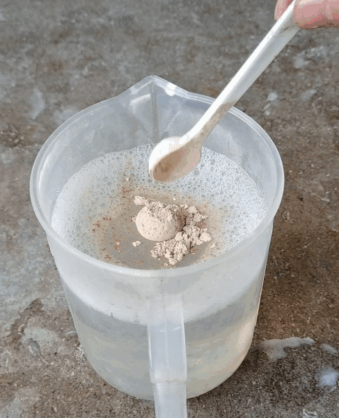 A spoonful of powder is being poured into a clear plastic measuring cup filled with water.